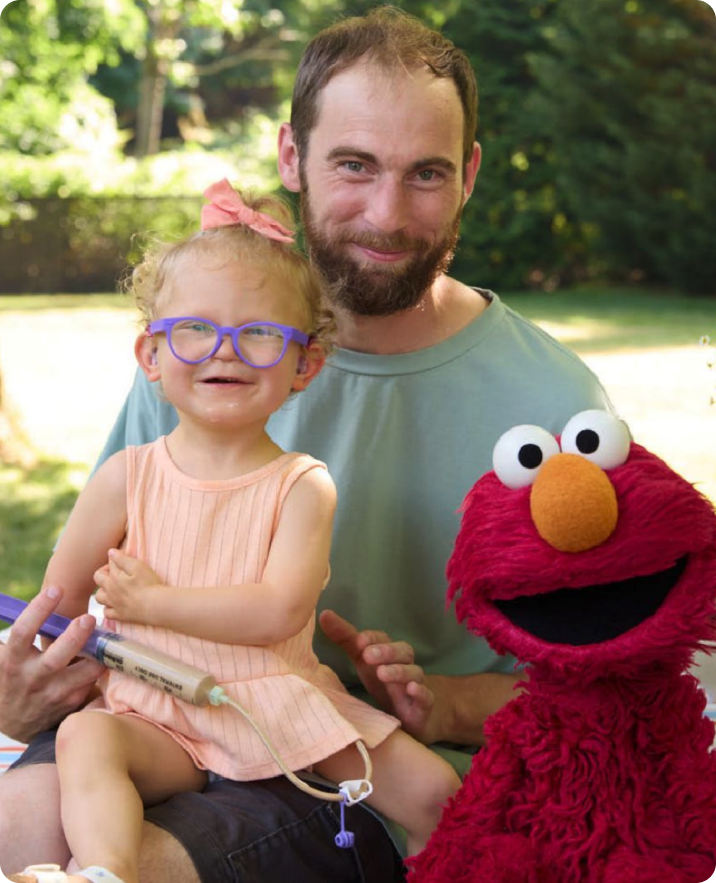 Father holding his daughter outdoors, with Elmo beside them